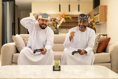 المشاركة والمنافسة بالألعاب التقنية الحديثة,  رجلان عربيان خليجيان عمانيان يرتديان الدشداشة والعمامة يجلسان على الاريكة في غرفة المعيشة يمسكان عصا التحكم, العالم الرقمي الترفيهي, المتعة والتسلية بالفيديو جيمز برفقة الأصدقاء، رفع قبضة اليد بايماءات السعادة والحماس