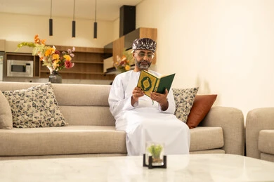 التقرب الى رب العالمين ، قراءة وحفظ القرآن الكريم، رجل عربي خليجي عماني يرتدي الدشداشة والعمامة يجلس على الاريكة في غرفة المعيشة يقوم بقراءة المصحف الشريف، تلاوة كتاب الله تعالى ,الاجتهاد و الخشوع في العباده