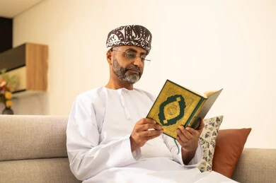 تلاوة كتاب الله تعالى , التقرب الى رب العالمين ، قراءة وحفظ القرآن الكريم، رجل عربي خليجي عماني يرتدي الدشداشة والعمامة يجلس على الاريكة في غرفة المعيشة يقوم بقراءة المصحف الشريف، الاجتهاد و الخشوع في العباده