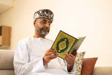 Drawing closer to the Lord of the worlds, reading and memorizing the Holy Quran, an Arab Gulf Omani man wearing a dishdasha and turban sits on the sofa in the living room reading the Holy Quran, reciting the Book of Allah, striving and being humble in worship.