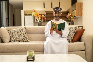 قراءة وحفظ القرآن الكريم، رجل عربي خليجي عماني يرتدي الدشداشة والعمامة يجلس على الاريكة في غرفة المعيشة يقوم بقراءة المصحف الشريف، تلاوة كتاب الله تعالى ,الاجتهاد و الخشوع في العباده, التقرب الى رب العالمين