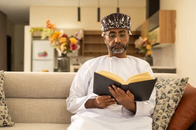 قراءة القصص والروايات الممتعة، مفهوم التثقف والتعلم الذاتي، الاستمتاع بالأجواء الهادئة، الشعور بالراحة والطمأنينة, رجل عربي خليجي عماني مبتسم يرتدي الدشداشة والعمامة يجلس على الاريكة في غرفة المعيشة يمسك بيده الكتاب، اتباع أسلوب حياة صحي