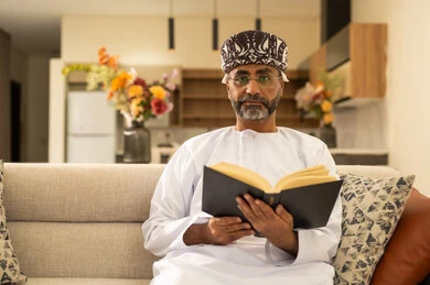 الشعور بالراحة والطمأنينة, قراءة القصص والروايات الممتعة، مفهوم التثقف والتعلم الذاتي، الاستمتاع بالأجواء الهادئة، رجل عربي خليجي عماني مبتسم يرتدي الدشداشة والعمامة يجلس على الاريكة في غرفة المعيشة يمسك بيده الكتاب، اتباع أسلوب حياة صحي