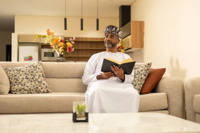 The concept of culture and self-learning, enjoying calm atmospheres, reading enjoyable stories and novels, feeling comfort and tranquility, a smiling Arab Gulf Omani man wearing a dishdasha and turban sitting on the sofa in the living room holding a book in his hand, following a healthy lifestyle.