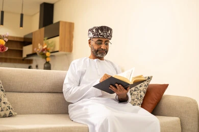 قراءة القصص والروايات الممتعة، مفهوم التثقف والتعلم الذاتي، الاستمتاع بالأجواء الهادئة، الشعور بالراحة والطمأنينة, رجل عربي خليجي عماني مبتسم يرتدي الدشداشة والعمامة يجلس على الاريكة في غرفة المعيشة يمسك بيده الكتاب، اتباع أسلوب حياة صحي