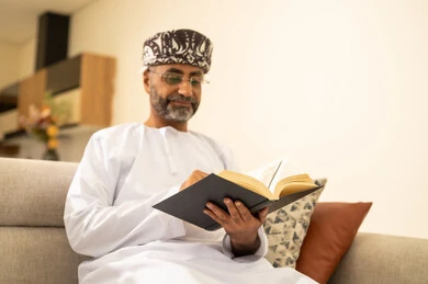 مفهوم التثقف والتعلم الذاتي، الاستمتاع بالأجواء الهادئة، قراءة القصص والروايات الممتعة، الشعور بالراحة والطمأنينة, رجل عربي خليجي عماني مبتسم يرتدي الدشداشة والعمامة يجلس على الاريكة في غرفة المعيشة يمسك بيده الكتاب، اتباع أسلوب حياة صحي