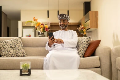 تصفح مواقع التواصل الاجتماعي عبر الانترنت، التواصل والدردشة مع الأصدقاء والأقارب عبر الجوال، قضاء وقت ممتع في المنزل، استخدام جهاز تقني حديث ومتطور، رجل عربي خليجي عماني يرتدي الدشداشة والعمامة يجلس في غرفة المعيشة يمسك بيده الهاتف المحمول