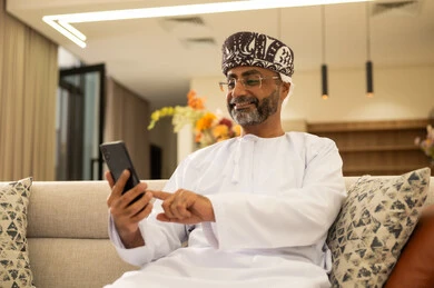 استخدام جهاز تقني حديث ومتطور، تصفح مواقع التواصل الاجتماعي عبر الانترنت، التواصل والدردشة مع الأصدقاء والأقارب عبر الجوال، قضاء وقت ممتع في المنزل، رجل عربي خليجي عماني يرتدي الدشداشة والعمامة يجلس في غرفة المعيشة يمسك بيده الهاتف المحمول استخدام جهاز تقني حديث ومتطور، تصفح مواقع التواصل الاجتماعي عبر الانترنت، التواصل والدردشة مع الأصدقاء والأقارب عبر الجوال، قضاء وقت ممتع في المنزل، رجل عربي خليجي عماني يرتدي الدشداشة والعمامة يجلس في غرفة المعيشة يمسك بيده الهاتف المحمول