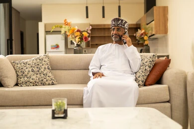 Gestures of joy and happiness, making a phone call, using a modern and advanced technical device, communicating and chatting with friends and relatives via mobile, an Arab Gulf Omani man wearing a dishdasha and a turban sitting in the living room holding a mobile phone in his hand.