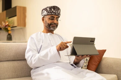 The use of modern technical and technological devices, communicating and chatting with friends and relatives via mobile, completing work tasks remotely at home, gestures of joy and happiness, an elderly Arab Gulf Omani man wearing a dishdasha and turban holding a mobile tablet sitting in the living room.
