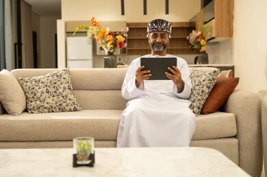 Completing remote work tasks at home, gestures of joy and happiness, using modern technological devices, communicating and chatting with friends and relatives via mobile, an elderly Arab Gulf Omani man wearing a dishdasha and turban holding a tablet in his hand sitting in the living room.