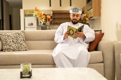 Drawing closer to the Lord of the worlds, reading and memorizing the Holy Quran, an Arab Gulf Omani man wearing a dishdasha and turban sits on the sofa in the living room reading the Holy Quran, reciting the Book of Allah, striving and being humble in worship.