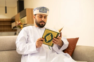 Drawing closer to the Lord of the worlds, reading and memorizing the Holy Quran, an Arab Gulf Omani man wearing a dishdasha and turban sits on the sofa in the living room reading the Holy Quran, reciting the Book of Allah, striving and being humble in worship.