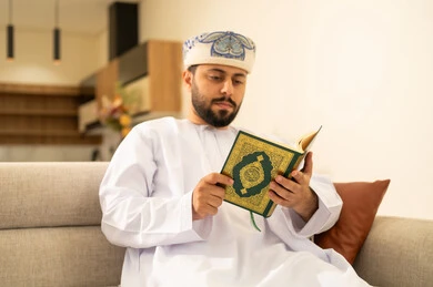Reflecting on the meanings of the Holy Quran, reciting the Book of Allah Almighty, drawing closer to the Lord of the Worlds, reading and memorizing the Holy Quran, an Arab Gulf Omani man wearing a dishdasha and turban sits on the sofa in the living room reading the Holy Quran, striving and being humble in worship.