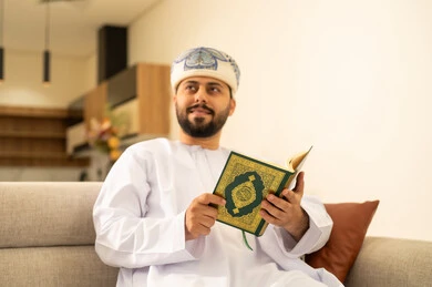قراءة وحفظ القرآن الكريم، رجل عربي خليجي عماني يرتدي الدشداشة والعمامة يجلس على الاريكة في غرفة المعيشة يقوم بقراءة المصحف الشريف، تلاوة كتاب الله تعالى ,الاجتهاد و الخشوع في العباده, التقرب الى رب العالمين