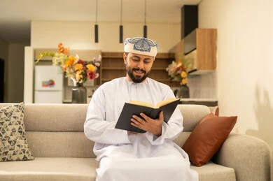 قراءة القصص والروايات الممتعة، مفهوم التثقف والتعلم الذاتي، الاستمتاع بالأجواء الهادئة، الشعور بالراحة والطمأنينة, رجل عربي خليجي عماني مبتسم يرتدي الدشداشة والعمامة يجلس على الاريكة في غرفة المعيشة يمسك بيده الكتاب، اتباع أسلوب حياة صحي