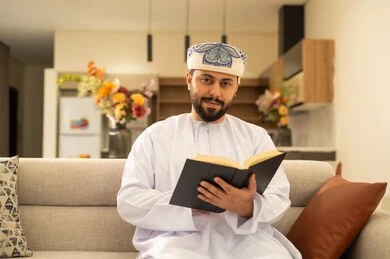 الشعور بالراحة والطمأنينة, قراءة القصص والروايات الممتعة، مفهوم التثقف والتعلم الذاتي، الاستمتاع بالأجواء الهادئة، رجل عربي خليجي عماني مبتسم يرتدي الدشداشة والعمامة يجلس على الاريكة في غرفة المعيشة يمسك بيده الكتاب، اتباع أسلوب حياة صحي