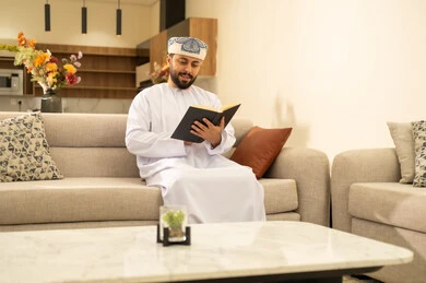 مفهوم التثقف والتعلم الذاتي، الاستمتاع بالأجواء الهادئة، قراءة القصص والروايات الممتعة، الشعور بالراحة والطمأنينة, رجل عربي خليجي عماني مبتسم يرتدي الدشداشة والعمامة يجلس على الاريكة في غرفة المعيشة يمسك بيده الكتاب، اتباع أسلوب حياة صحي