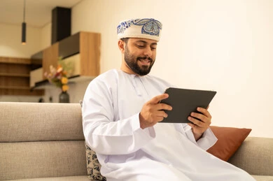 Spending enjoyable times at home, gestures of joy and happiness, using modern technological devices, communicating and chatting with friends and relatives via mobile, an Arab Gulf Omani man wearing a dishdasha and turban holding a tablet in his hand sitting in the living room.