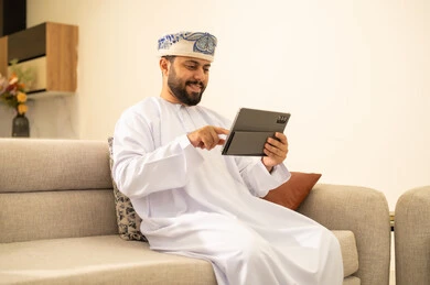 Gestures of joy and happiness, using modern technological devices, communicating and chatting with friends and relatives via mobile, spending enjoyable times at home, an Arab Gulf Omani man wearing a dishdasha and turban holding a tablet in his hand sitting in the living room.