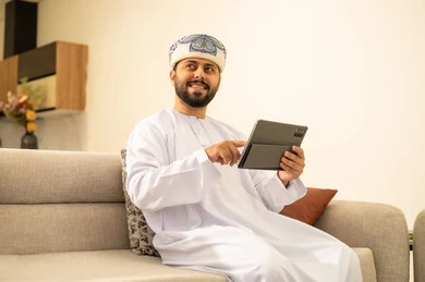Expressions of joy and happiness, using modern technological devices, communicating and chatting with friends and relatives via mobile, spending enjoyable times at home, an Arab Gulf Omani man wearing a dishdasha and turban holding a tablet in his hand sitting in the living room.
