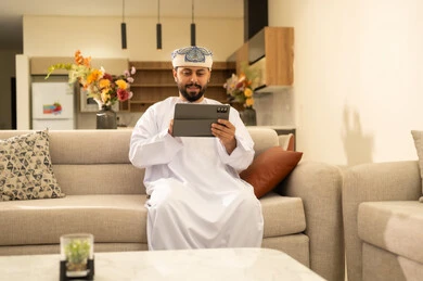 ايماءات الفرح والسعادة، استخدام الأجهزة التقنية والتكنولوجية الحديثة، التواصل والدردشة مع الأصدقاء والأقارب عبر الجوال، قضاء أوقات ممتعة في المنزل, رجل عربي خليجي عماني يرتدي الدشداشة والعمامة يمسك بيده التابليت المحمول يجلس في غرفة المعيشة