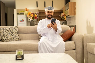 قضاء أوقات ممتعة في المنزل, استخدام الأجهزة التقنية والتكنولوجية الحديثة، التواصل والدردشة مع الأصدقاء والأقارب عبر الجوال، تعابير وايماءات الفرح والسعادة، رجل عربي خليجي عماني يرتدي الدشداشة والعمامة يمسك بيده الهاتف المحمول يجلس في غرفة المعيشة قضاء أوقات ممتعة في المنزل, استخدام الأجهزة التقنية والتكنولوجية الحديثة، التواصل والدردشة مع الأصدقاء والأقارب عبر الجوال، تعابير وايماءات الفرح والسعادة، رجل عربي خليجي عماني يرتدي الدشداشة والعمامة يمسك بيده الهاتف المحمول يجلس في غرفة المعيشة