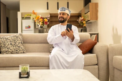 التواصل والدردشة مع الأصدقاء والأقارب عبر الجوال، قضاء أوقات ممتعة في المنزل, استخدام الأجهزة التقنية والتكنولوجية الحديثة، ايماءات الفرح والسعادة، رجل عربي خليجي عماني يرتدي الدشداشة والعمامة يمسك بيده الهاتف المحمول يجلس في غرفة المعيشة