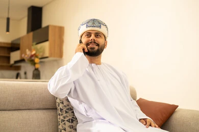 Making a phone call, expressions of joy and happiness, using modern technological devices, communicating and chatting with friends and relatives via mobile, spending enjoyable times at home, an Arab Gulf Omani man wearing a dishdasha and turban holding a mobile phone sitting in the living room.