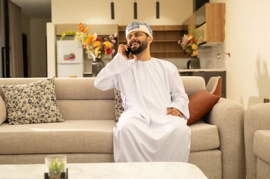 إجراء مكالمة هاتفية، تعابير الفرح والسعادة، استخدام الأجهزة التقنية والتكنولوجية الحديثة، التواصل والدردشة مع الأصدقاء والأقارب عبر الجوال، قضاء أوقات ممتعة في المنزل, رجل عربي خليجي عماني يرتدي الدشداشة والعمامة يمسك بيده الهاتف المحمول يجلس في غرفة المعيشة