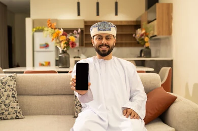 A blank black screen displayed on the mobile phone with expressions of joy and happiness, using modern technological devices, communicating and chatting with friends and relatives via mobile, spending enjoyable times at home, an Arab Gulf Omani man wearing a dishdasha and turban holding a mobile phone sitting in the living room.