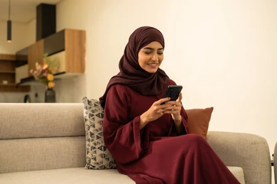 Watching favorite movies and series, following social media sites online, completing work tasks remotely, an Omani woman using modern and advanced devices and technologies, a Saudi Gulf Arab woman sitting on a sofa in the living room holding a mobile phone in her hand.