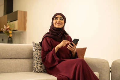 Completing work tasks remotely, watching favorite movies and series, following social media sites online, an Omani woman using modern and advanced devices and technologies, a Saudi Gulf Arab woman sitting on a sofa in the living room holding a mobile phone in her hand.