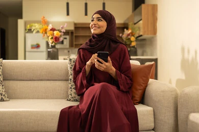 متابعة مواقع التواصل الاجتماعي عبر الانترنت، القيام بإنجاز مهام العمل عن بعد, مشاهدة الأفلام والمسلسلات المفضلة، سيدة عمانية تقوم باستخدام الأجهزة والتقنيات الحديثة والمتطورة, امرأة عربية خليجية سعودية تجلس على أريكة في غرفة المعيشة و تمسك بيدها الهاتف المحمول