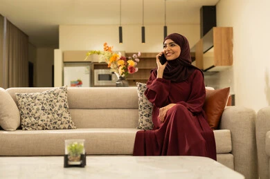 Making a phone call via mobile, communicating with family and friends, completing work tasks remotely, an Omani woman using modern devices and technologies with expressions of joy, a Saudi Gulf Arab woman sitting on a sofa in the living room holding a mobile phone in her hand.