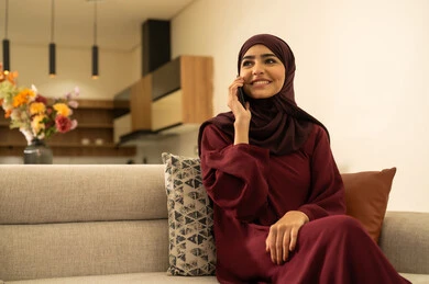 Communicating with family and friends, making a phone call via mobile, completing work tasks remotely, an Omani woman using modern devices and technologies with expressions of joy, a Saudi Arabian Gulf Arab woman wearing a hijab and an abaya sitting on a sofa in the living room holding a mobile phone in her hand.