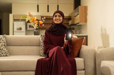 عرض شاشة سوداء فارغة عبر الجوال، متابعة مواقع التواصل الاجتماعي عبر الانترنت، القيام بإنجاز مهام العمل عن بعد، سيدة عمانية تقوم باستخدام الأجهزة والتقنيات الحديثة بتعابير السرور, امرأة عربية خليجية سعودية تجلس على أريكة في غرفة المعيشة و تمسك بيدها الهاتف المحمول