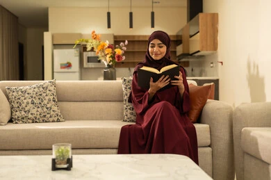 Relaxing and enjoying the calm atmosphere, feeling comfortable and at peace, an Omani woman is reading enjoyable novels and stories, a smiling Saudi Gulf Arab woman wearing an abaya and hijab sitting on the sofa in the living room holding a book in her hand, following a healthy lifestyle.