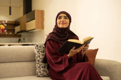 الشعور بالراحة والطمأنينة, امرأة عمانية تقوم بقراءة الروايات والقصص الممتعة، سيدة عربية خليجية سعودية مبتسمة ترتدي عباءة وحجاب تجلس على الاريكة في غرفة المعيشة تمسك بيدها الكتاب، الاسترخاء والاستمتاع بالأجواء الهادئة، اتباع أسلوب حياة صحي