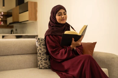 الشعور بالراحة والطمأنينة, امرأة عمانية تقوم بقراءة الروايات والقصص الممتعة، سيدة عربية خليجية سعودية مبتسمة ترتدي عباءة وحجاب تجلس على الاريكة في غرفة المعيشة تمسك بيدها الكتاب، الاسترخاء والاستمتاع بالأجواء الهادئة، اتباع أسلوب حياة صحي
