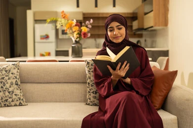 الاسترخاء والاستمتاع بالأجواء الهادئة، الشعور بالراحة والطمأنينة, امرأة عمانية تقوم بقراءة الروايات والقصص الممتعة، سيدة عربية خليجية سعودية مبتسمة ترتدي عباءة وحجاب تجلس على الاريكة في غرفة المعيشة تمسك بيدها الكتاب، اتباع أسلوب حياة صحي