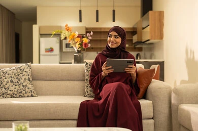 متابعة مواقع التواصل الاجتماعي عبر الانترنت، القيام بإنجاز مهام العمل عن بعد, مشاهدة الأفلام والمسلسلات المفضلة، سيدة عمانية تقوم باستخدام الأجهزة والتقنيات الحديثة والمتطورة, امرأة عربية خليجية سعودية تجلس على أريكة في غرفة المعيشة و تمسك بيدها جهاز التابليت