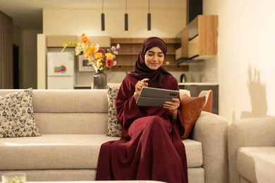 مشاهدة الأفلام والمسلسلات المفضلة، متابعة مواقع التواصل الاجتماعي عبر الانترنت، القيام بإنجاز مهام العمل عن بعد, سيدة عمانية تقوم باستخدام الأجهزة والتقنيات الحديثة والمتطورة, امرأة عربية خليجية سعودية تجلس على أريكة في غرفة المعيشة و تمسك بيدها جهاز التابليت