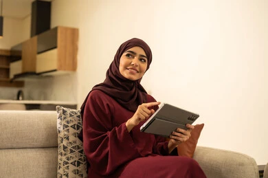 القيام بإنجاز مهام العمل عن بعد, مشاهدة الأفلام والمسلسلات المفضلة، متابعة مواقع التواصل الاجتماعي عبر الانترنت، سيدة عمانية تقوم باستخدام الأجهزة والتقنيات الحديثة والمتطورة, امرأة عربية خليجية سعودية تجلس على أريكة في غرفة المعيشة و تمسك بيدها جهاز التابليت