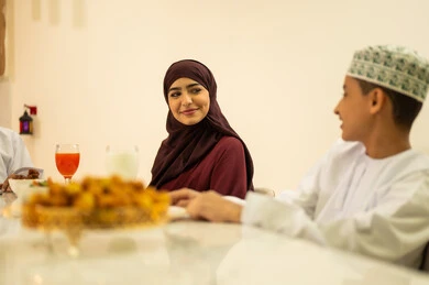 The month of goodness and blessings, the celebration of the arrival of the blessed month of Ramadan, family visits and gatherings during Islamic holidays and occasions, an Arab Gulf Omani boy wearing a dishdasha and kumma sits at the iftar table in Ramadan with his family, enjoying the sweet dish of luqaimat.