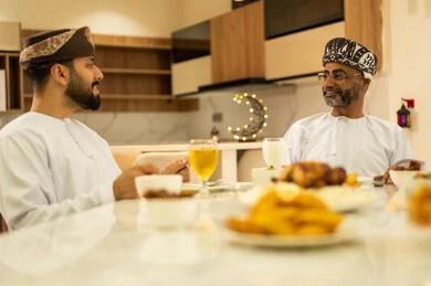 اجتماع العائلة على المائدة، سفرة مليئة بالمقبلات والاطباق الرمضانية الشهية، لمة الاصدقاء على سفرة رمضان, صديقان عربيان خليجيان عمانيان يرتديان الدشداشة والعمامة يقومان بتناول الطعام بايماءات السرور بقدوم الشهر الفضيل, الاحتفال بالاعياد و المناسبات الاسلامية
