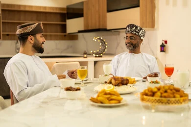 A table filled with appetizers and delicious Ramadan dishes, a gathering of friends at the Ramadan table, two Arab Gulf friends from Oman wearing the dishdasha and turban enjoying the food with gestures of joy at the arrival of the holy month, celebrating Islamic holidays and occasions, family gathering at the table.