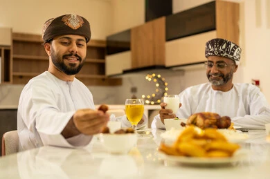 اجتماع العائلة على المائدة، سفرة مليئة بالمقبلات والاطباق الرمضانية الشهية، لمة الاصدقاء على سفرة رمضان, صديقان عربيان خليجيان عمانيان يرتديان الدشداشة والعمامة يقومان بتناول الطعام بايماءات السرور بقدوم الشهر الفضيل, الاحتفال بالاعياد و المناسبات الاسلامية