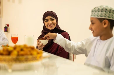 The month of goodness and blessings, the celebration of the arrival of the blessed month of Ramadan, family visits and gatherings during Islamic holidays and occasions, an Arab Gulf Omani boy wearing a dishdasha and kumma sits at the iftar table in Ramadan with his family, enjoying the sweet dish of luqaimat.