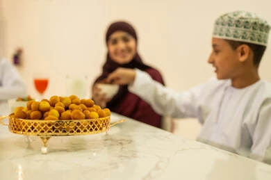 شهر الخير والبركة ، الاحتفال بقدوم شهر رمضان المبارك ، الزيارات والتجمعات الأسريه في الاعياد والمناسبات الاسلامية ، ولد عربي خليجي عماني يرتدي الدشداشة والكمة يجلس على مائدة الافطار فى شهر رمضان مع عائلته ، تناول حلى اللقيمات .